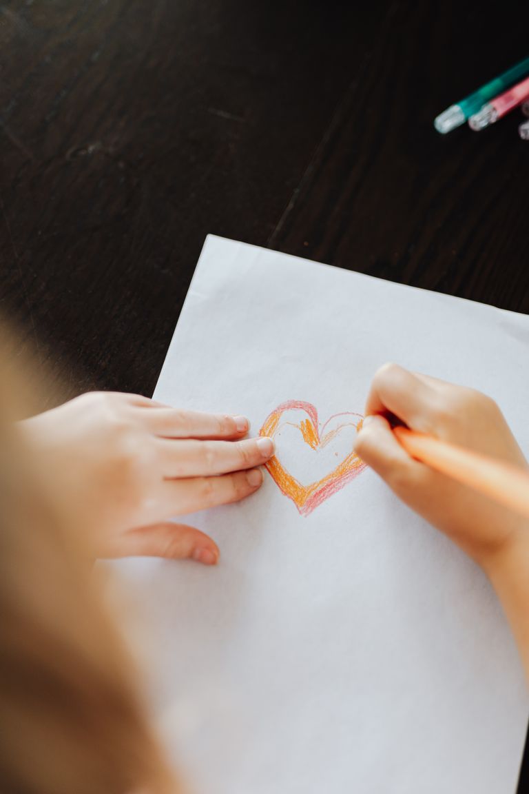 A child draws a colorful heart on white paper using colored pencils.