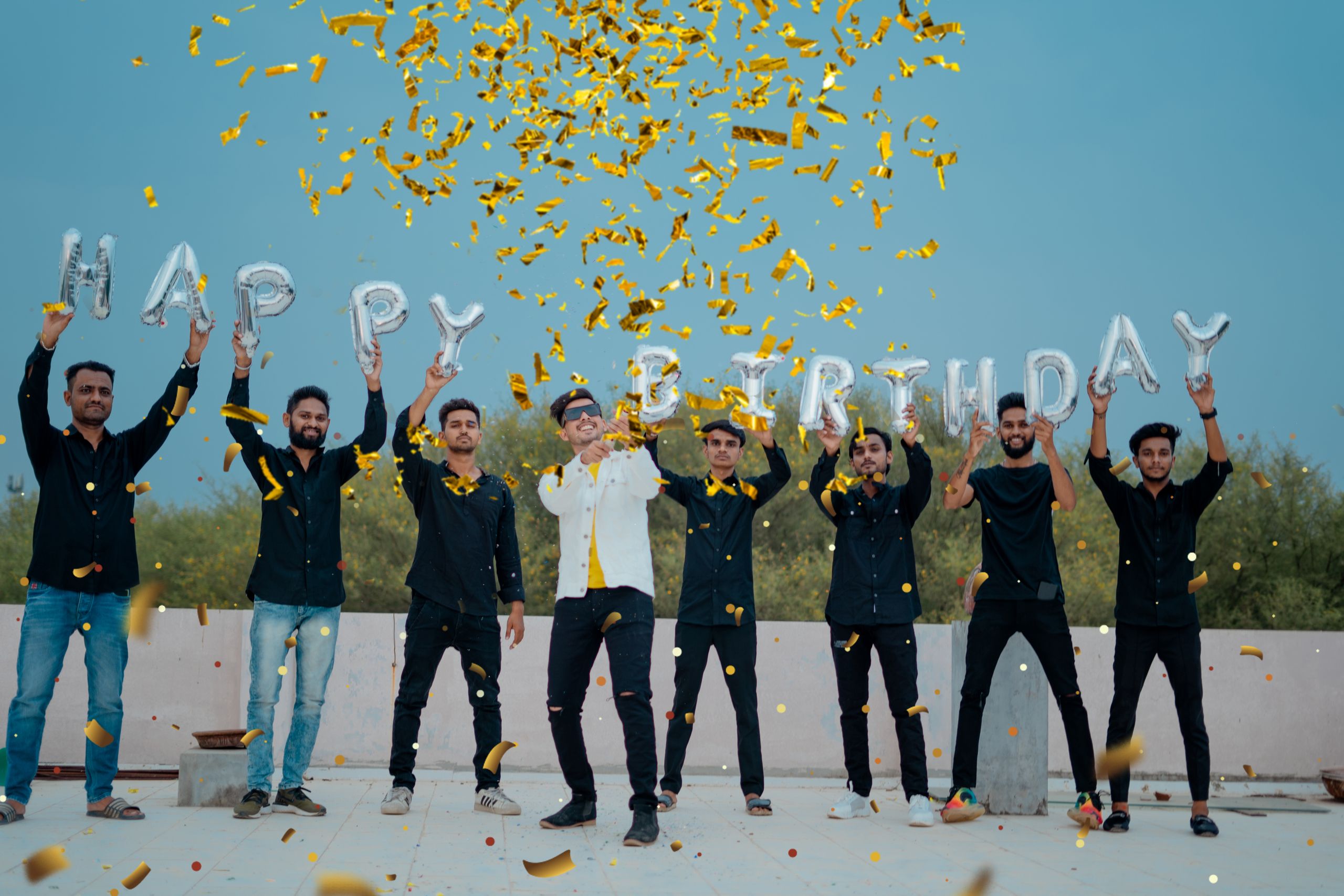 A joyful group of young men celebrates a birthday outdoors with confetti and balloon letters.