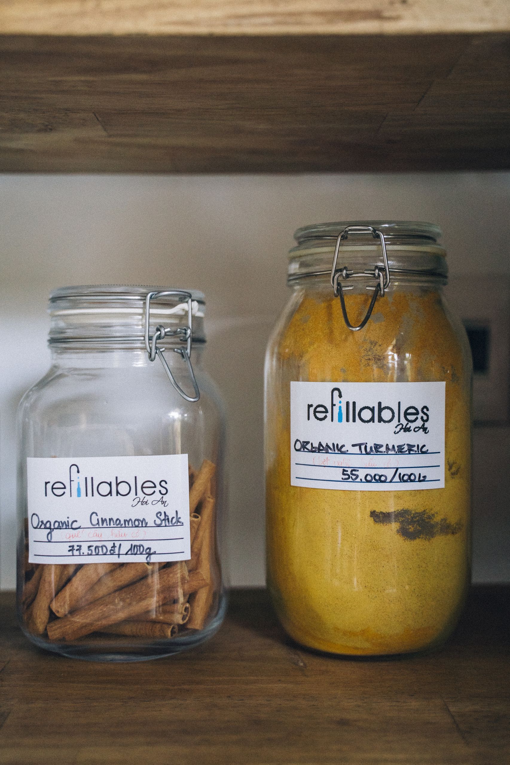 Close-up of reusable glass jars with organic cinnamon sticks and turmeric labeled on a wooden shelf.