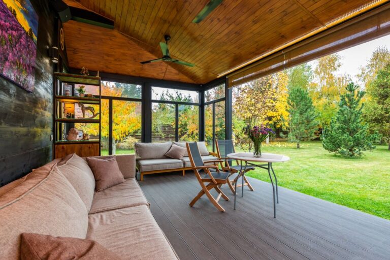 Sofas placed near wooden wall decorated with picture veranda furnished with armchairs near table under fan on ceiling Stock Photo.