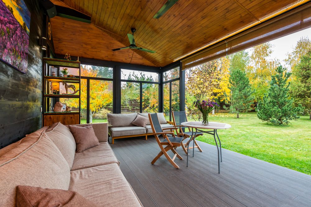 Sofas placed near wooden wall decorated with picture veranda furnished with armchairs near table under fan on ceiling Stock Photo.