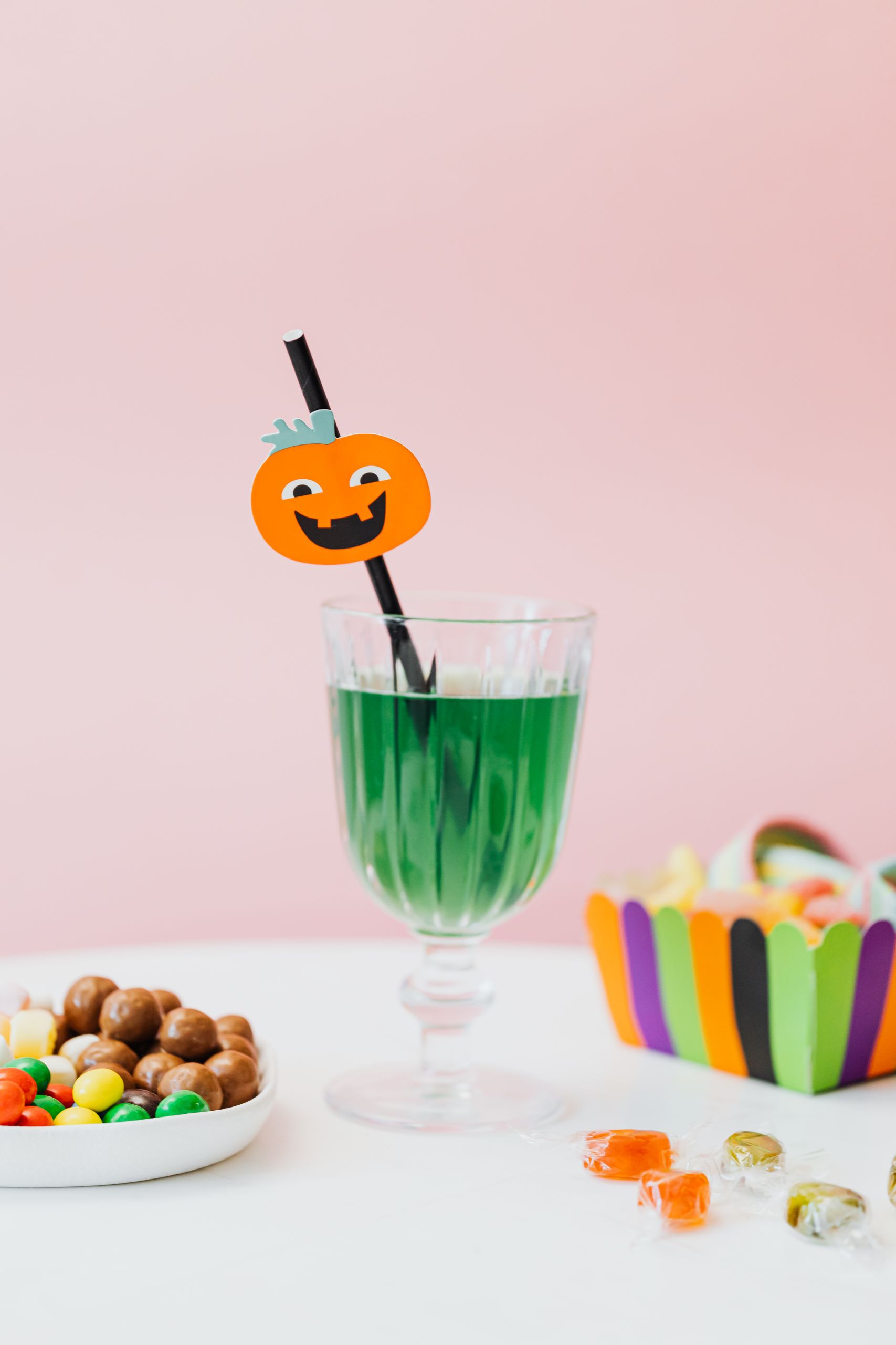 Festive Halloween green drink with a pumpkin-decorated straw, surrounded by colorful candies on a pink backdrop.