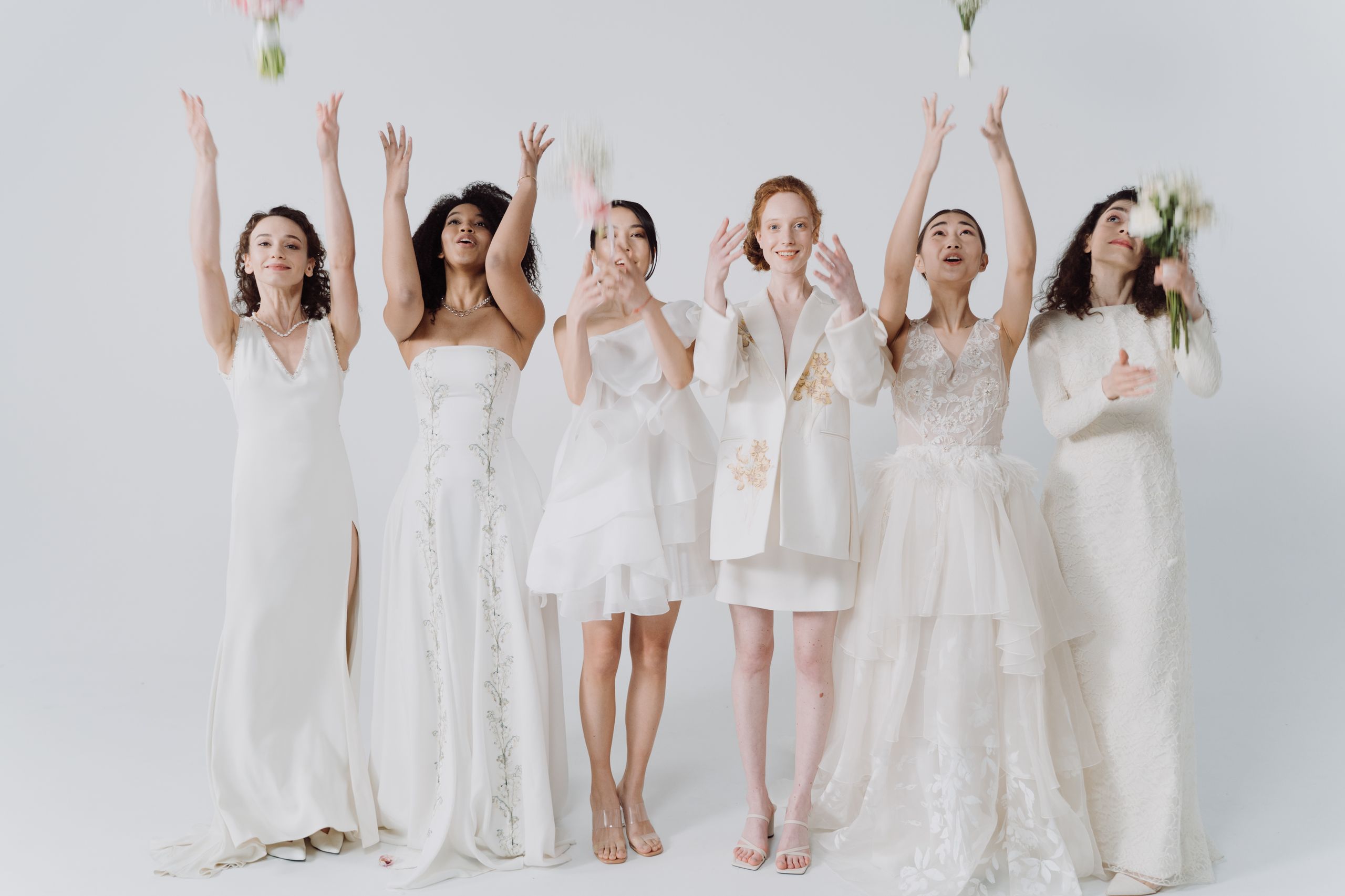 Joyful group of diverse women celebrating in elegant white dresses, throwing bouquets.