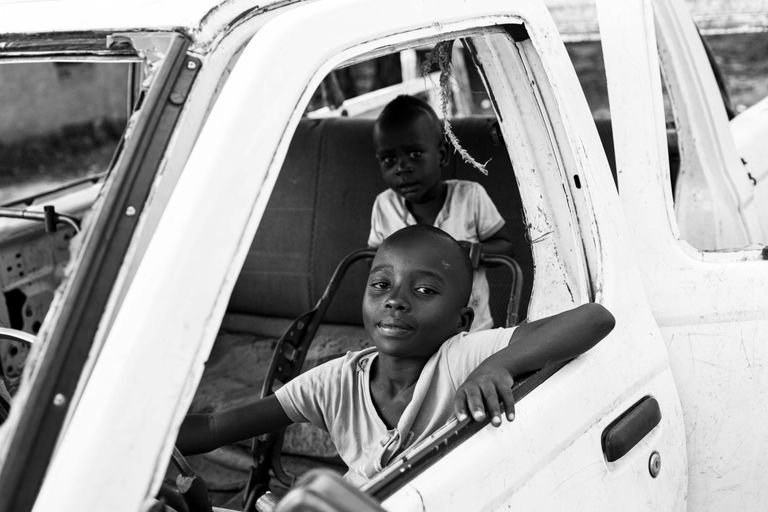 Two African children playing in an old car, captured in black and white.