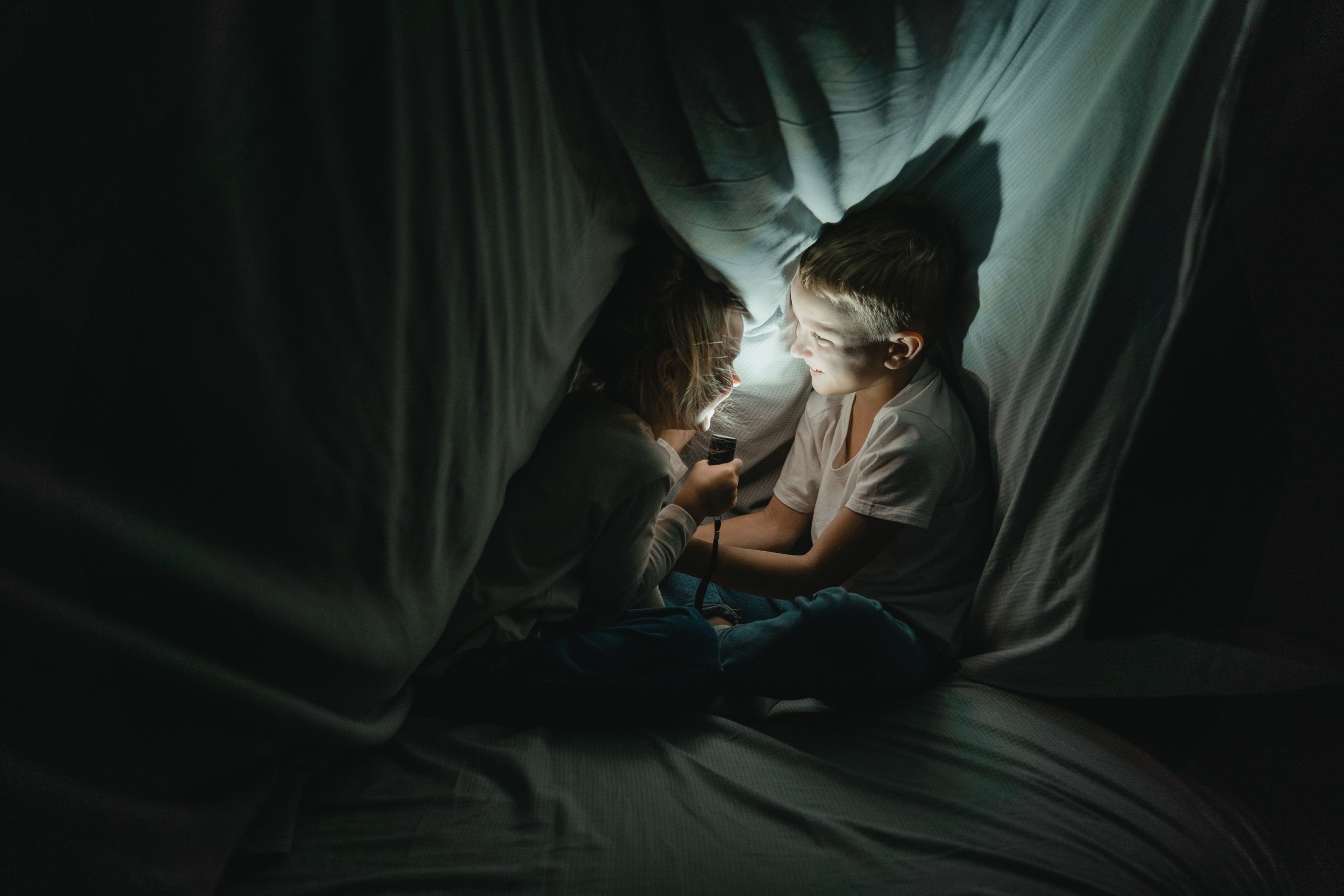 Two kids enjoy storytelling under a cozy blanket fort lit by a flashlight.